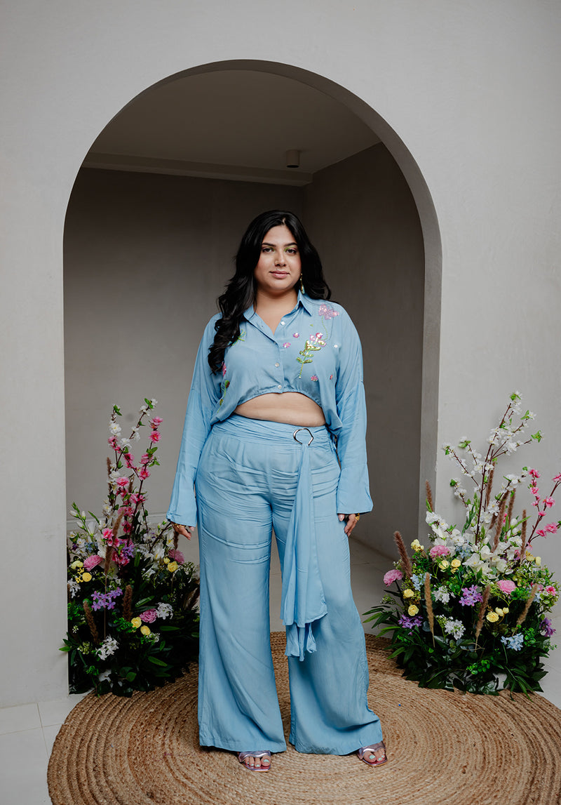 Model wearing Balmy Coord Set, a sky-blue floral hand-embroidered co-ord with bell sleeves and straight-fit pants, standing in a modern archway.